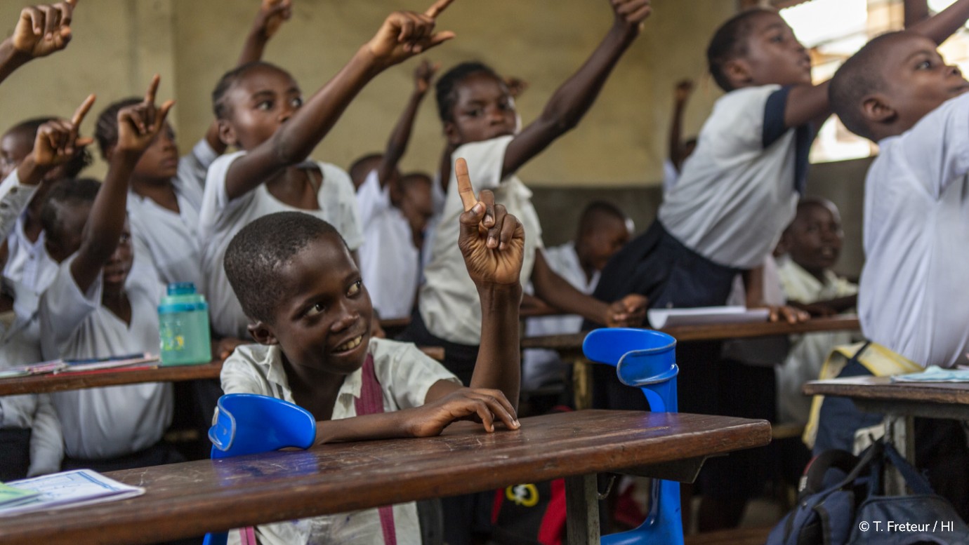 Dans une salle de classe à Kinshasa en RDC, au premier plan un enfant assis avec des béquilles de chaque côté lève le doigt. Au second plan, plusieurs de ces camarades font de même.