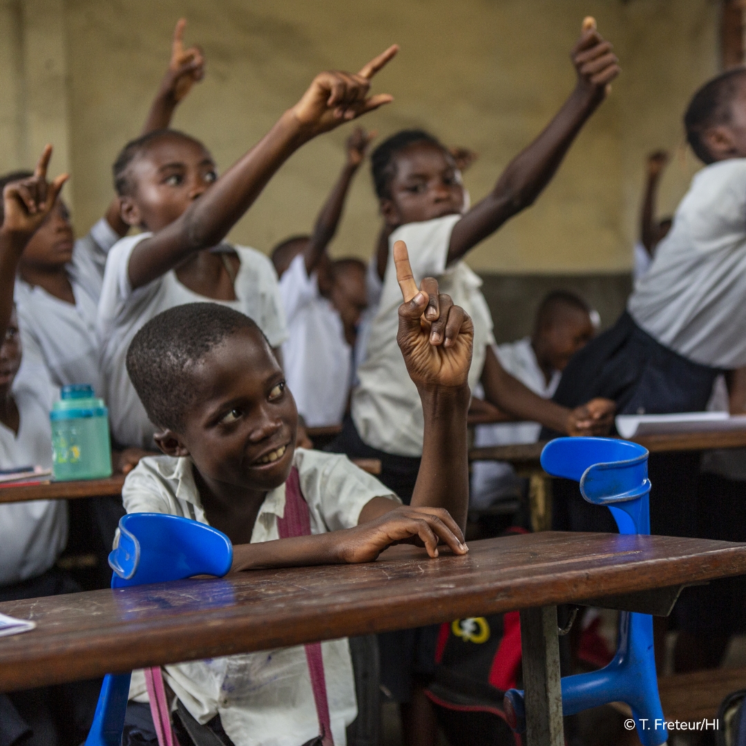 Dans une salle de classe à Kinshasa en RDC, au premier plan un enfant assis avec des béquilles de chaque côté lève le doigt. Au second plan, plusieurs de ces camarades font de même.