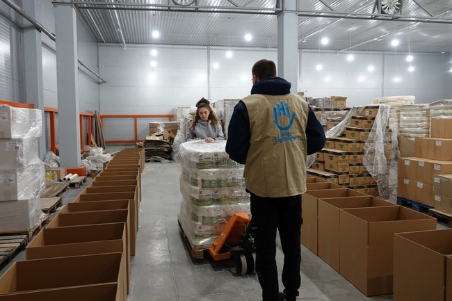 Dans un entrepôt rempli de cartons, deux personnes manipulent des palettes.