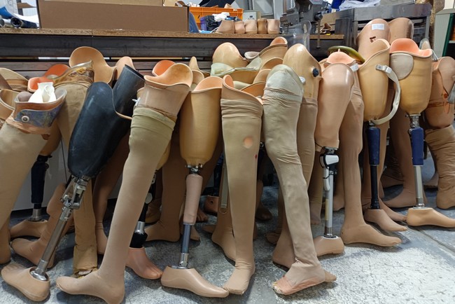 Dans un atelier, plusieurs prothèses de jambe sont alignées posées contre un établi.