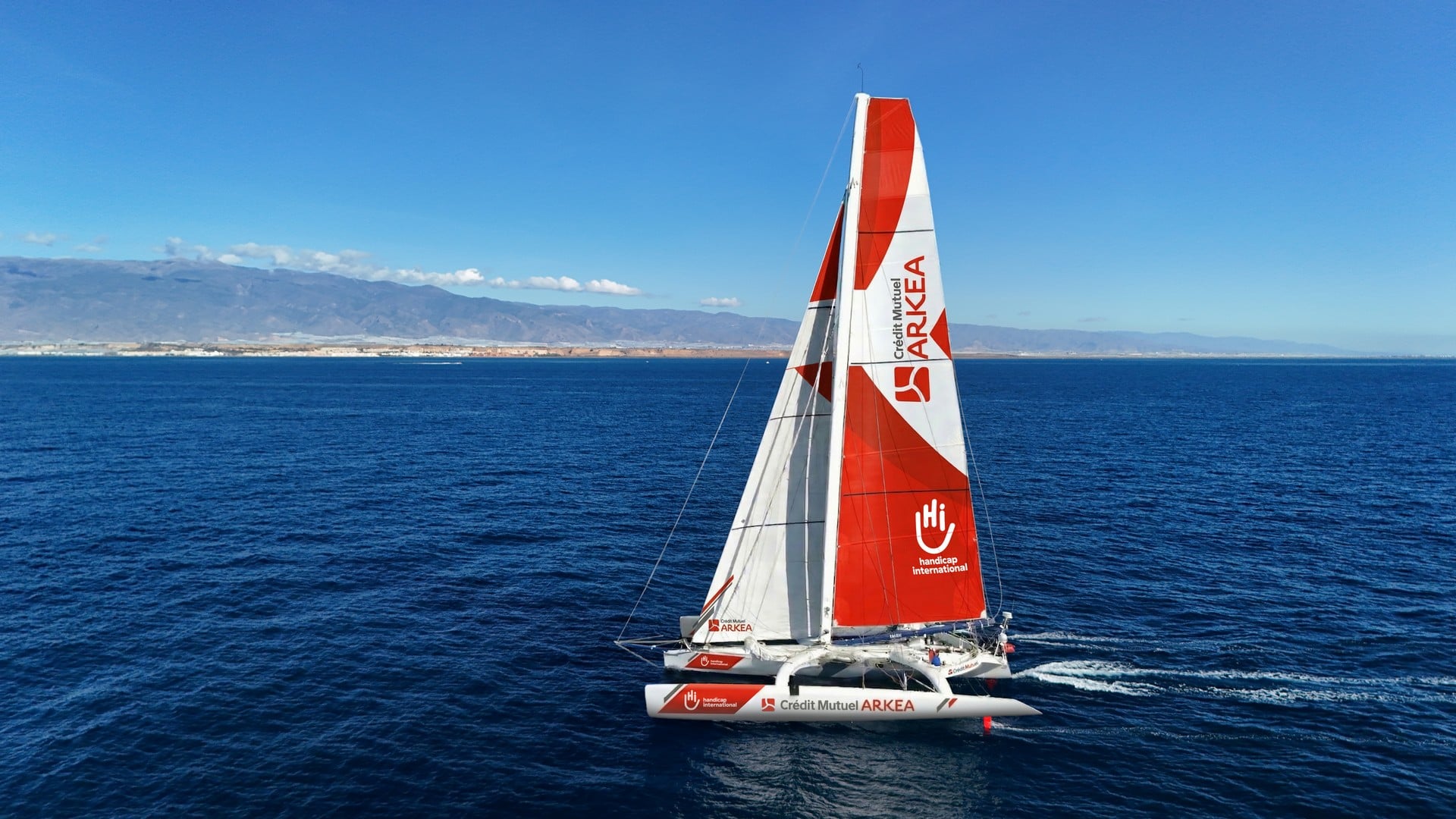 Un trimaran rouge et blanc navigue sur la mer près des côtes.