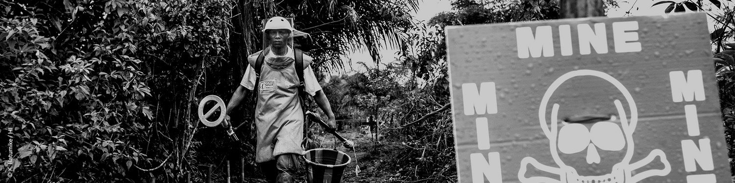 Photo en noir et blanc montrant un homme en tenue de démineur à l'orée d'un bois et qui regarde l'objectif. Sur la droite au premier plan, on devine un panneau montrant une tête de mort avec le mot MINE sur chacun des quatre côté du panneau.