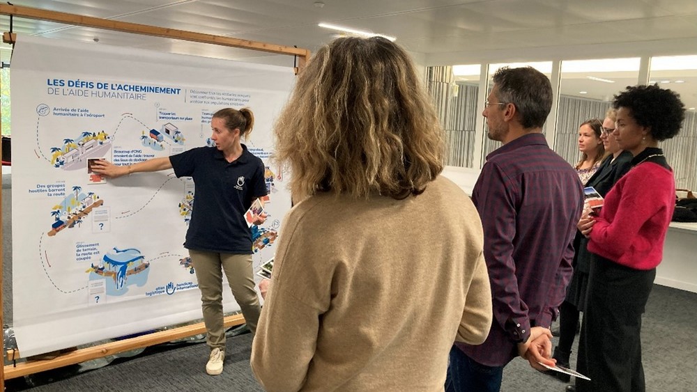 Cinq personnes debout dans une pièce assistent à une présentation par une sixième qui leur montre des dessins sur un panneau.