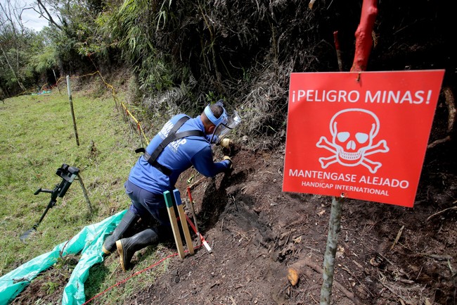 Sur un terrain accidenté, un démineur en tenue est à genoux en train de travailler. Sur la droite, un panneau rouge indiquant un danger lié aux mines.