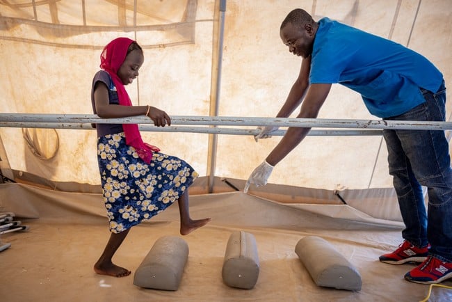 Au Tchad, une jeune fille s'entraîne à marcher sur un parcours de réadaptation, encouragée par un personnel humanitaire.