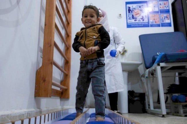 Un jeune garçon se tient debout dans une salle de rééducation et sourit au photographe.