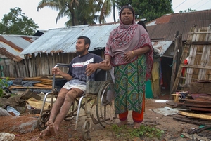 Devant des maisons touchées par des inondations au Bangladesh, un homme en fauteuil roulant et une femme à ses côtés.