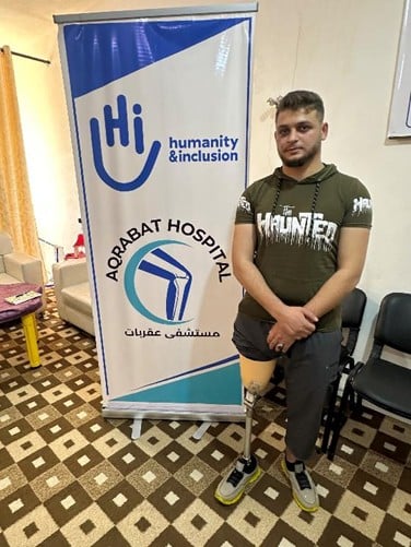 Mohammad, un jeune Syrien, pose dans une pièce à côté d'un kakémono avec les logos de l'hôpital d'Aqrabat et de HI.