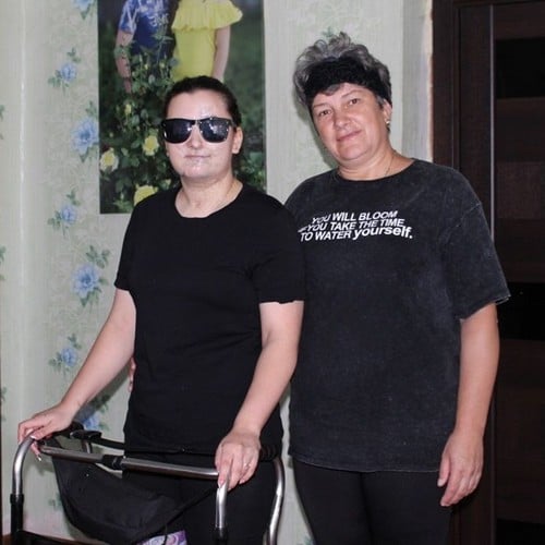 Une femme aveugle équipée de lunettes noires se tient debout dans une pièce à l'aide d'un déambulateur, une autre femme se tient à ses côtés.