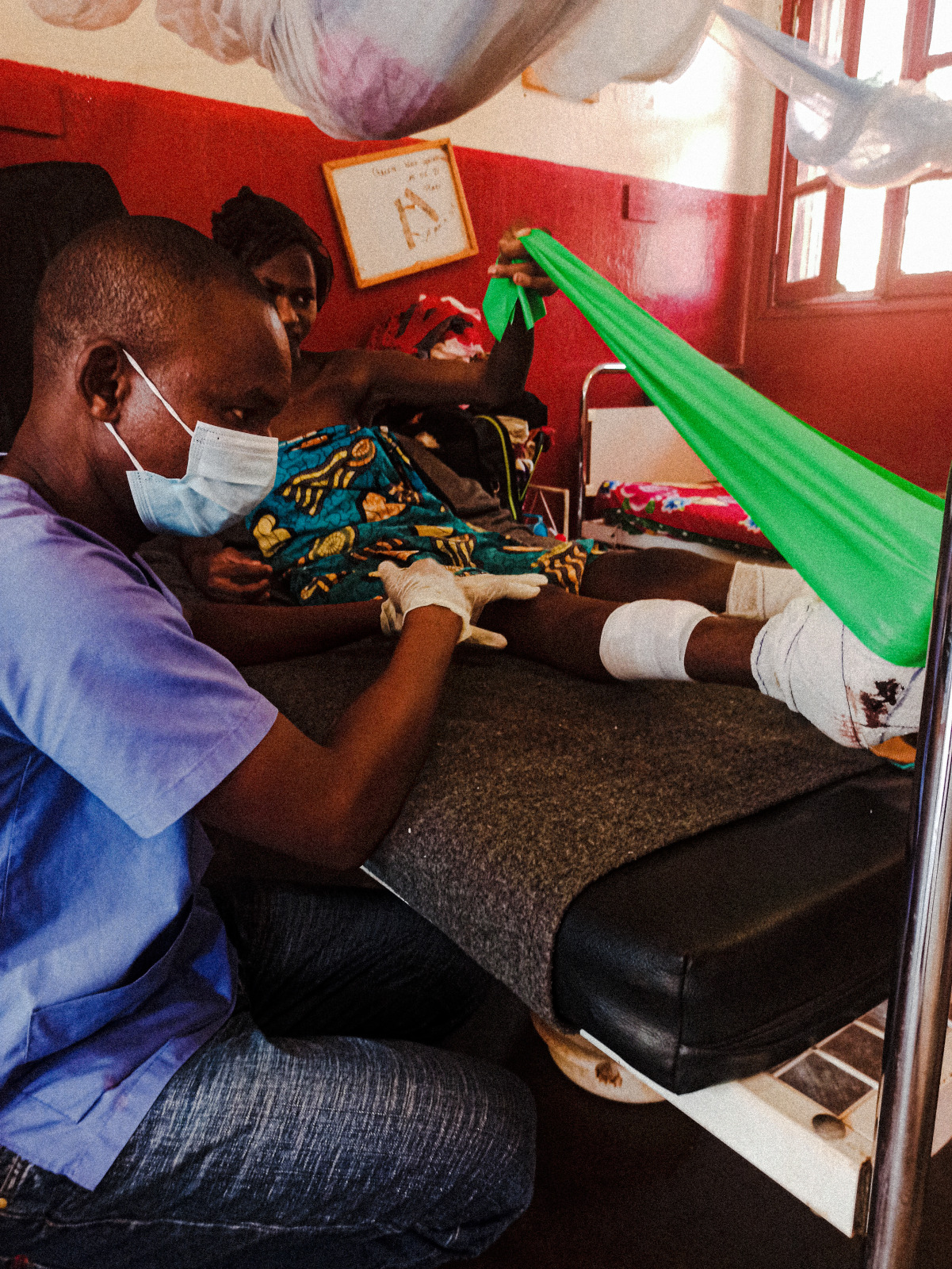 Un kinésithérapeute de Handicap International aide Alima à faire des exercices pour ses jambes depuis son lit à l'hôpital - © A. Servant / HI