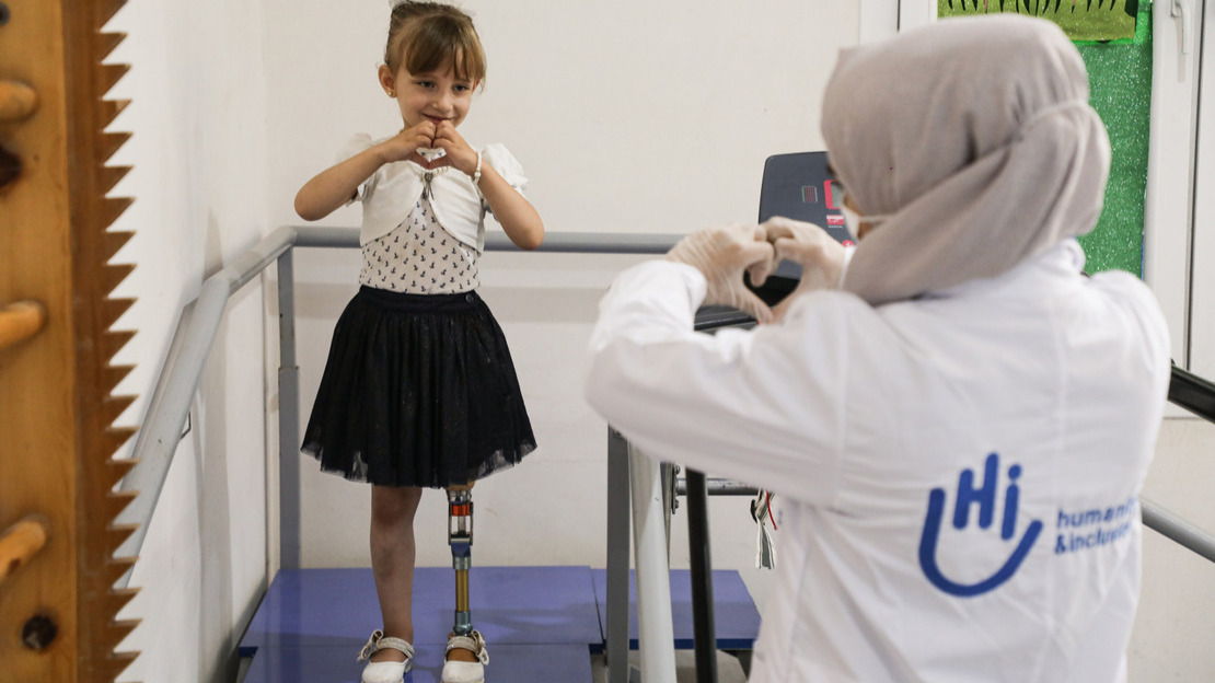 Photo d'Enas, une petite fille de 5 ans qui porte une jupe noire avec un haut et un gilet blanc. Elle est debout sur un escalier de rééducation avec des rampes. On voit ses jambes, dont sa prothèse. Enas regarde Fatima, la travailleuse de HI en face d'elle. Enas et Fatima font un cœur avec leurs mains.