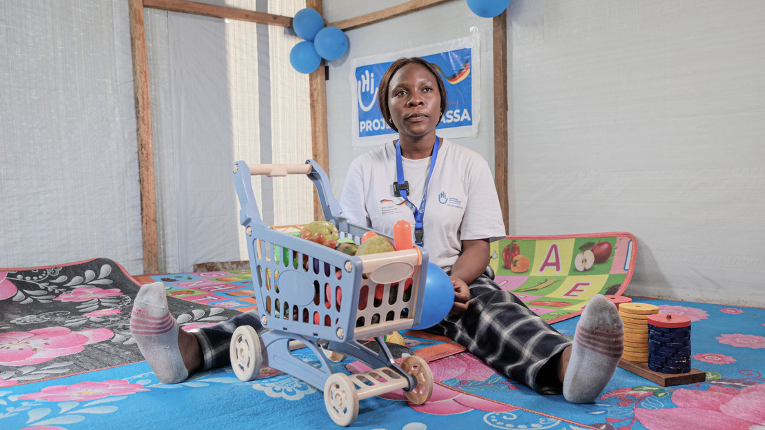 Une kinésithérapeute est assise dans une salle. Elle tient un ballon dans les mains et autour d'elles sont disposés des jeux d'éveil pour enfants.