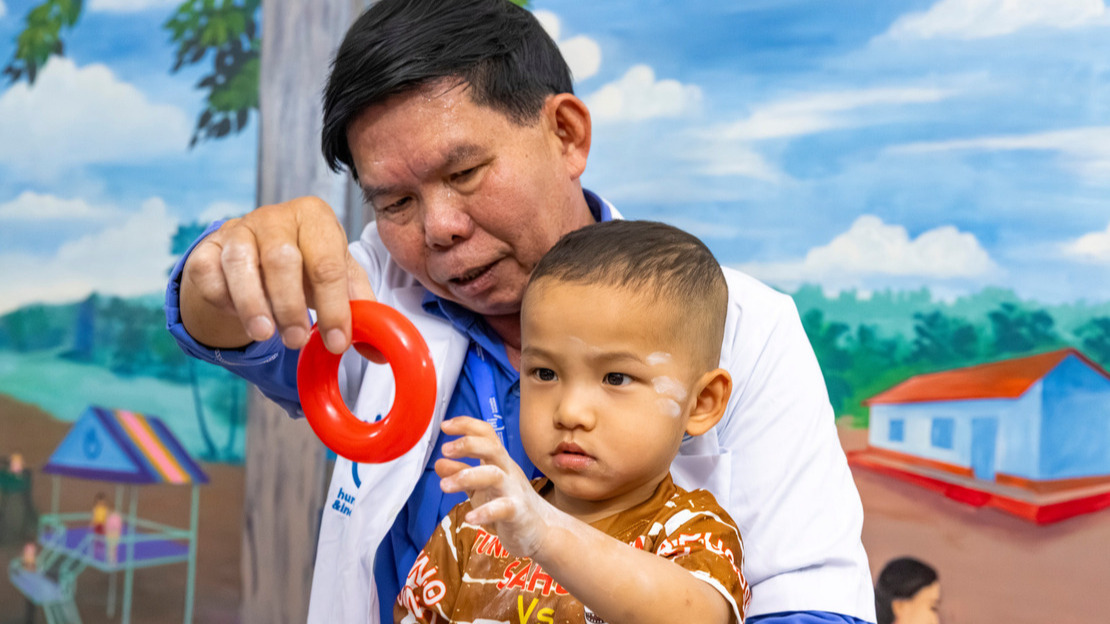 Portrait en gros plan d'un homme en blouse blanche qui tient sur ses genoux un jeune garçon. Le garçon tend la main pour attraper un petit anneau en plastique que l'homme tient devant lui.
