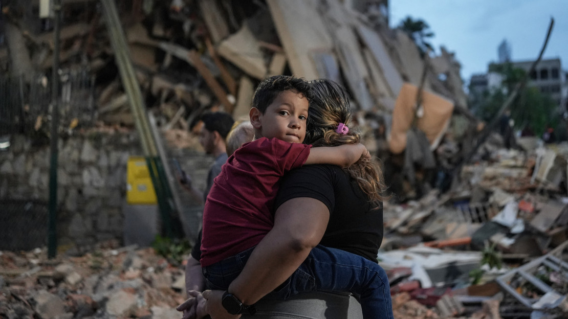 Au premier plan, une femme de dos porte dans ses bras un enfant qui regarde fixement le photographe. Derrière eux, on voit des bâtiments effondrés.