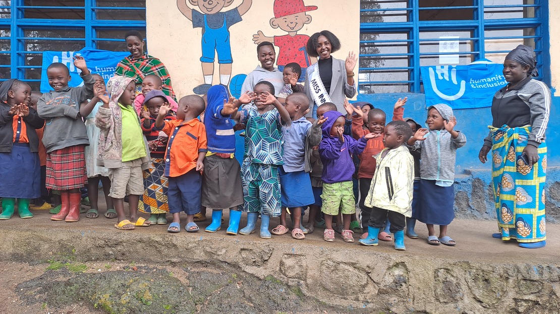 Un groupe d'enfants sourit et fait signe à la caméra. Derrière eux, Miss Jeannette salue également en souriant. Ils se tiennent tous devant un mur d'école décoré par des dessins.