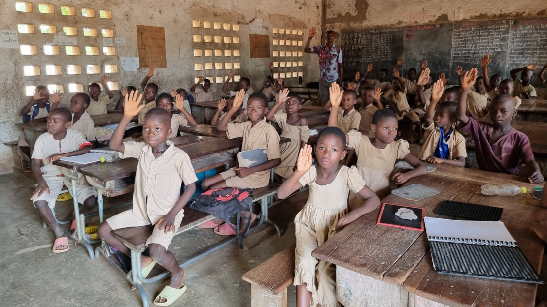 Intérieur d'une salle de classe. Des enfants en uniformes beige âgés de 8 à 10 ans sont assis autour de pupitres en bois et lèvent la main.