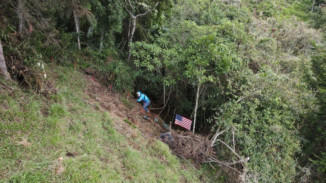 Un démineur est accroupi sur une pente raide et démine le terrain devant lui. Autour de lui, une forêt luxuriante. Un drapeau des États-Unis figure à quelques mètres de lui.