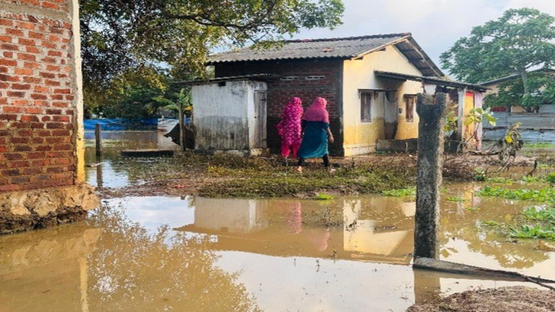 Au Sri Lanka, deux femmes vêtues de vêtements colorés marchent dans une zone inondée, à proximité de maisons en briques et en béton. Le sol est recouvert d'une eau boueuse.