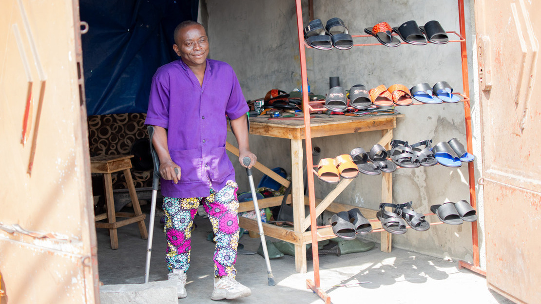 Un homme se tient debout, appuyé sur des béquilles, entre les portes ouvertes d'un atelier donnant sur la rue. Un présentoir expose différentes paires de chaussures de type sandales.