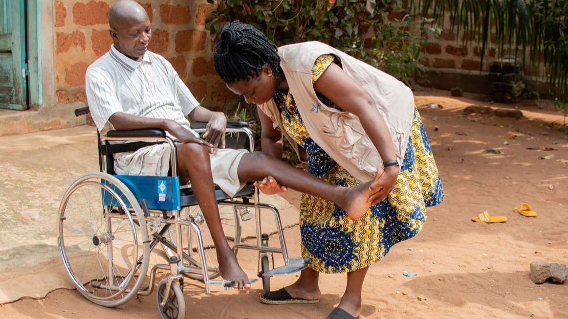Un homme assis dans un fauteuil roulant regarde le mouvement effectué par une femme debout à ses côtés et qui tend sa jambe gauche.