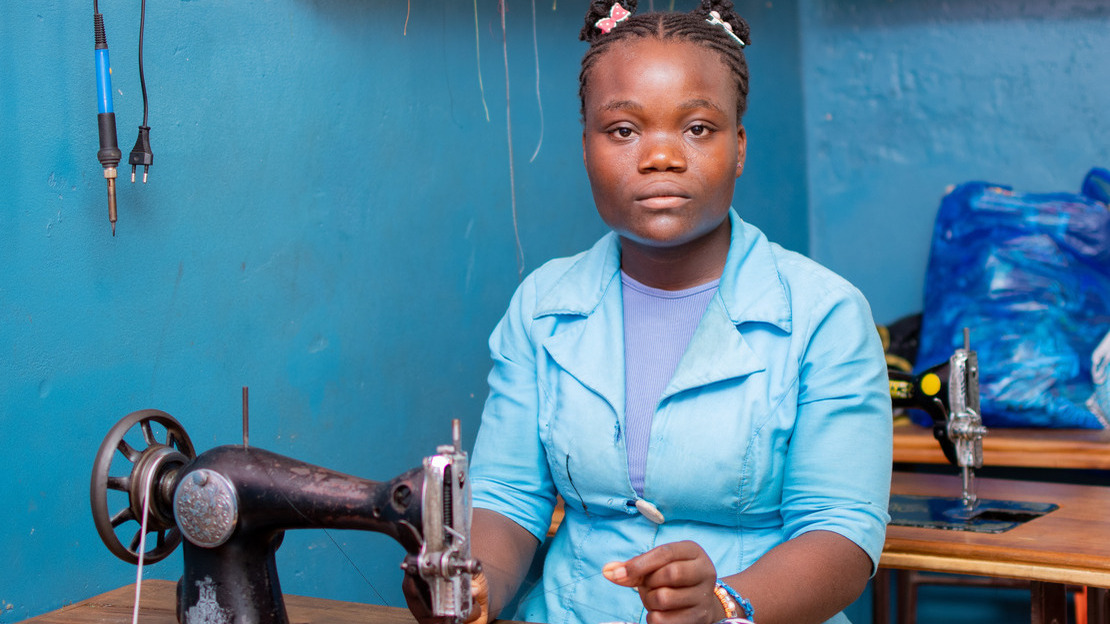 Portrait d'une adolescente assise face à une table et qui regarde le photographe. Devant elle est posée une machine à coudre et un morceau de tissu. Elle tire un fil passé dans la machine.