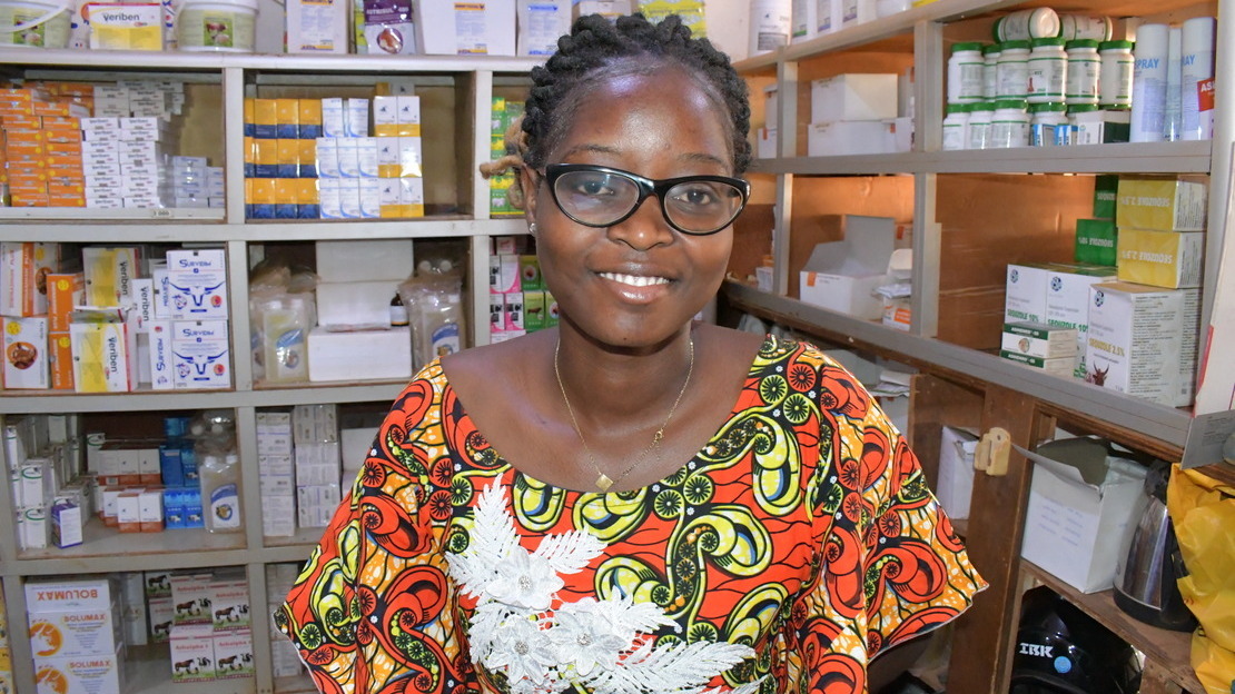 Portrait d'une jeune femme dans une pharmacie, avec des rayonnages remplis de boîtes de médicaments. La jeune femme porte des lunettes et sourit.