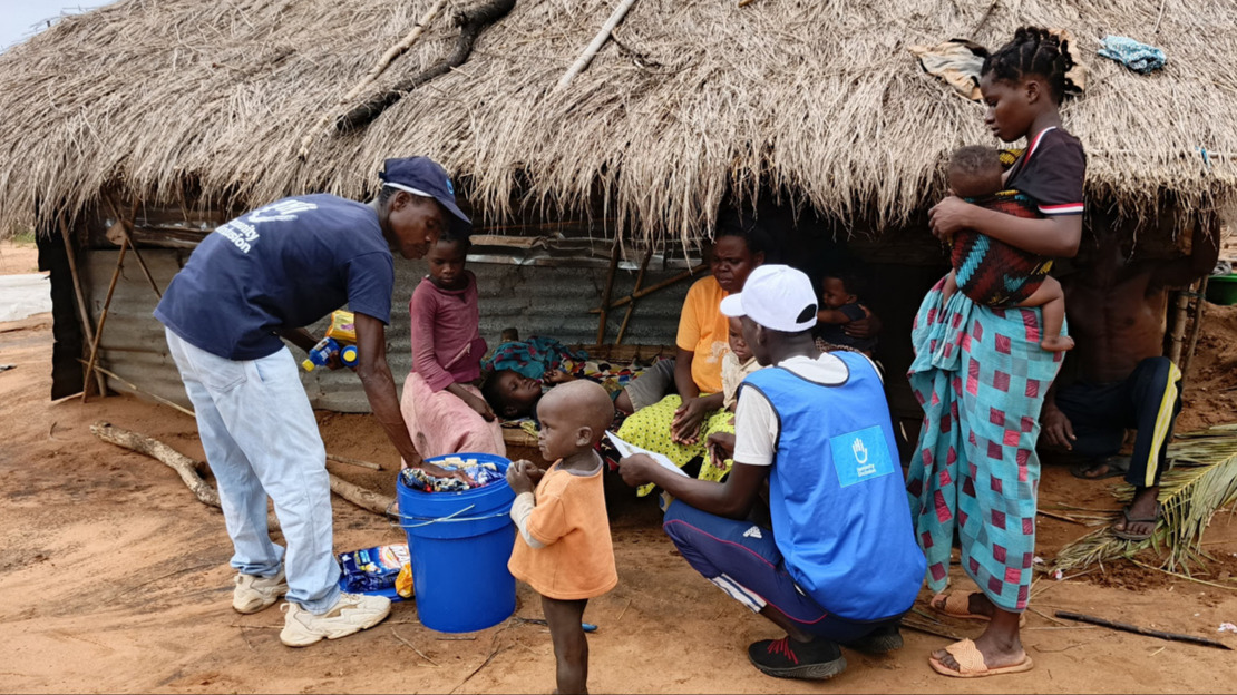 Devant une case en tôle et en paille, deux personnes portant des t-shirt floqués HI déballent le contenu d'un grand seau bleu en montrant ce qu'il contient à une famille composée de deux femmes et de plusieurs enfants en bas âge.