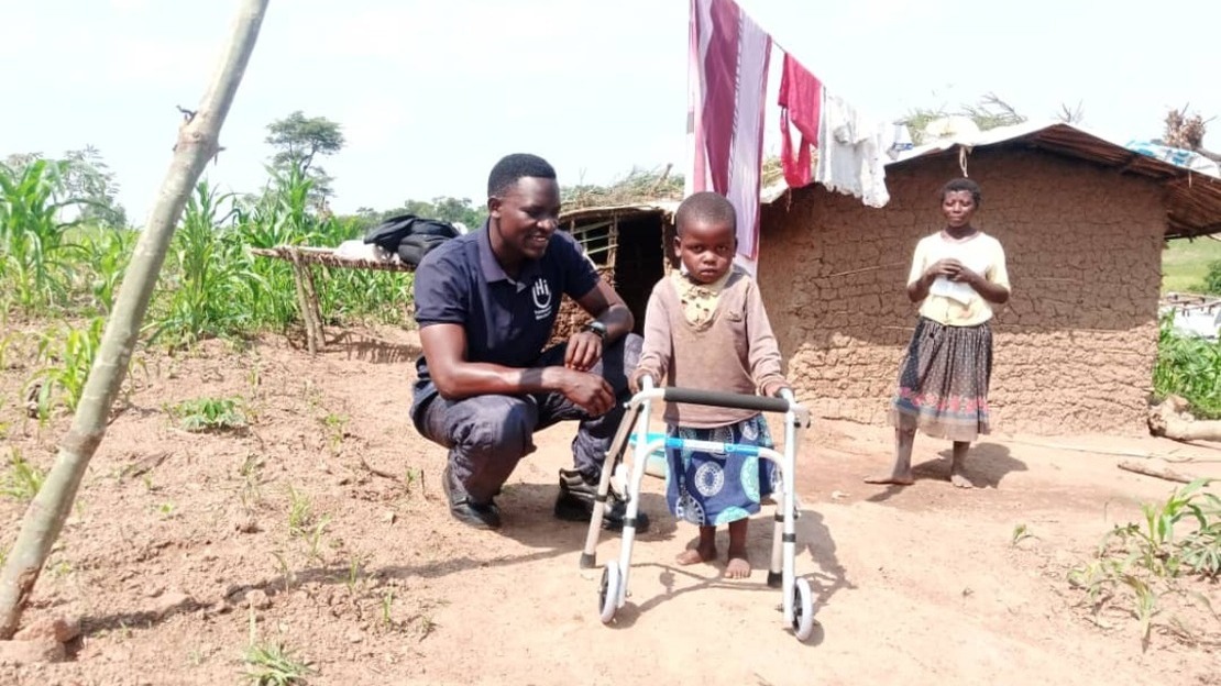 Devant une maison en briques, une petite fille se tient appuyée sur un déambulateur. À genoux à côté d'elle, un homme sourit. Une femme se tient debout à l'arrière-plan.