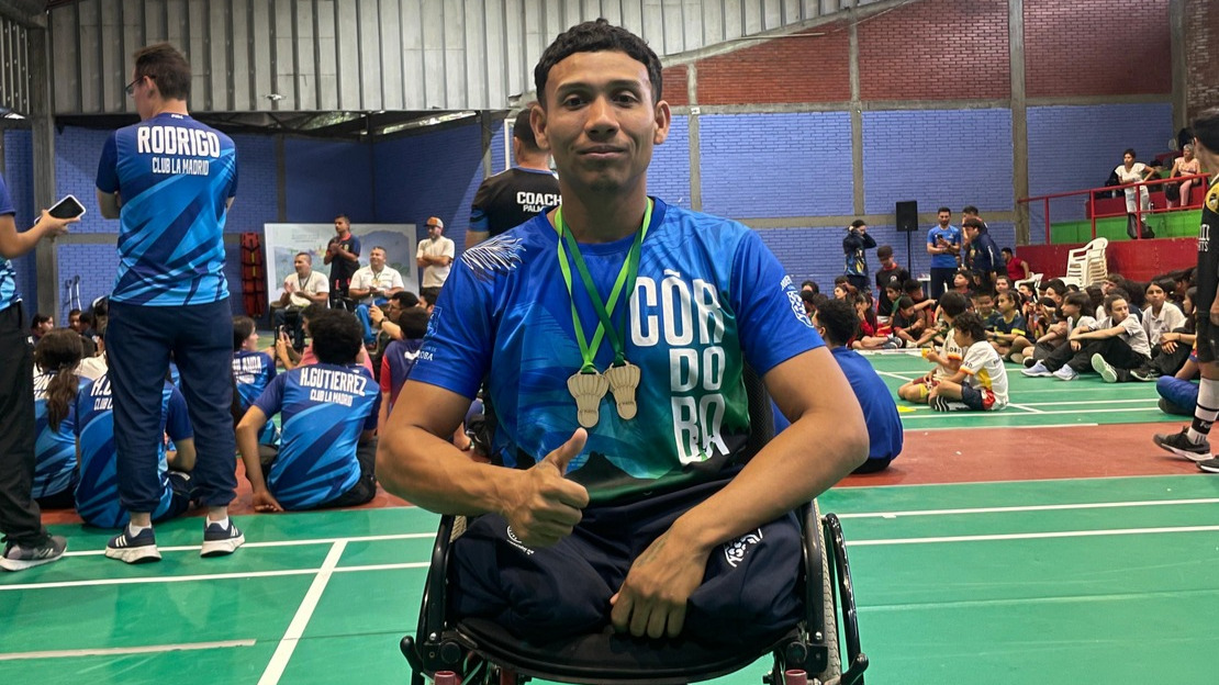 Un jeune homme en fauteuil roulant porte un maillot sportif et a deux médailles passées autour du cou. Derrière lui, on devine un stade couvert.