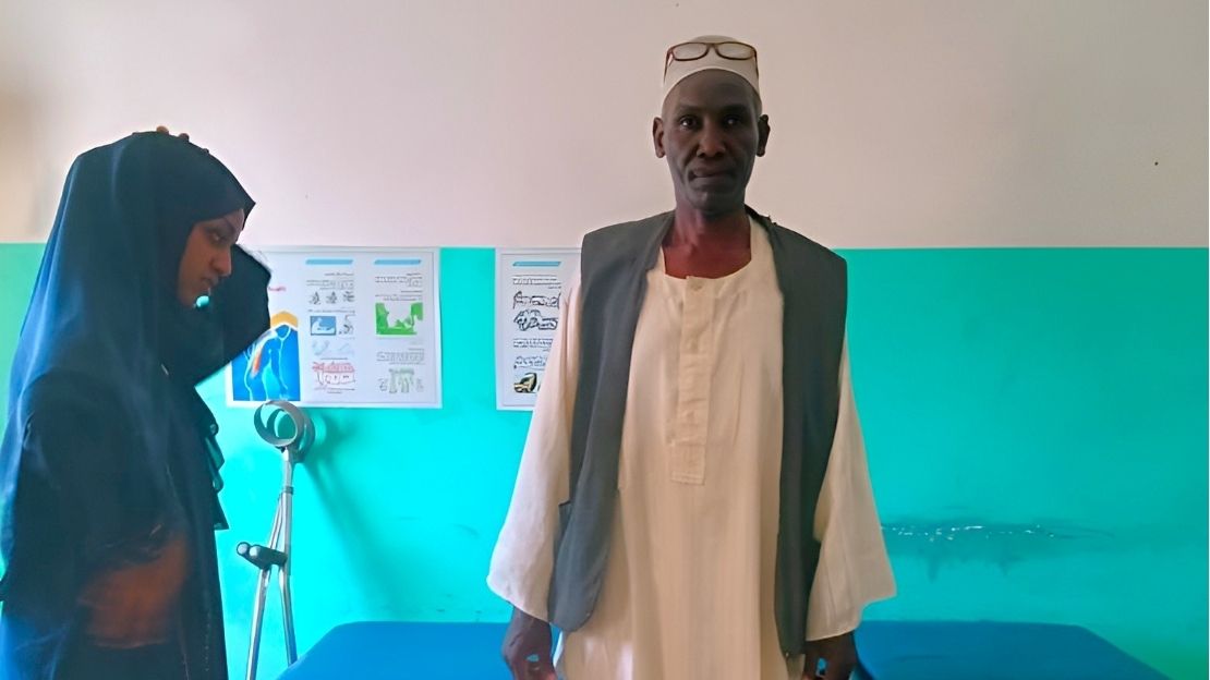 Un homme se tient debout et regarde le photographe. À l'arrière-plan, quelques béquilles sont appuyées contre le mur.