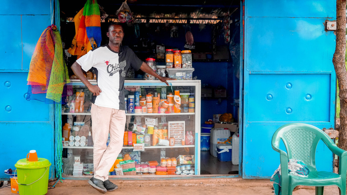 Un homme s'appuie sur un comptoir en verre derrière lequel sont exposé de nombreux produits (d'hygiène, alimentaire, d'entretien...). Des deux côtés, des portes métalliques bleues sont grand ouvertes sur l'intérieur de la boutique.