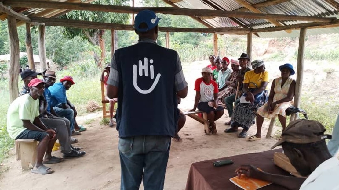 Sous un abri de tôle, Un homme se tient de dos et porte un t-shirt avec le logo HI. Face et autour de lui, assis sur des bancs, des hommes et des femmes l'écoutent.