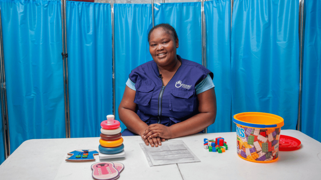 Une femme souriante est assise derrière un bureau sur lequel sont posés des jouets et matériels de rééducation.