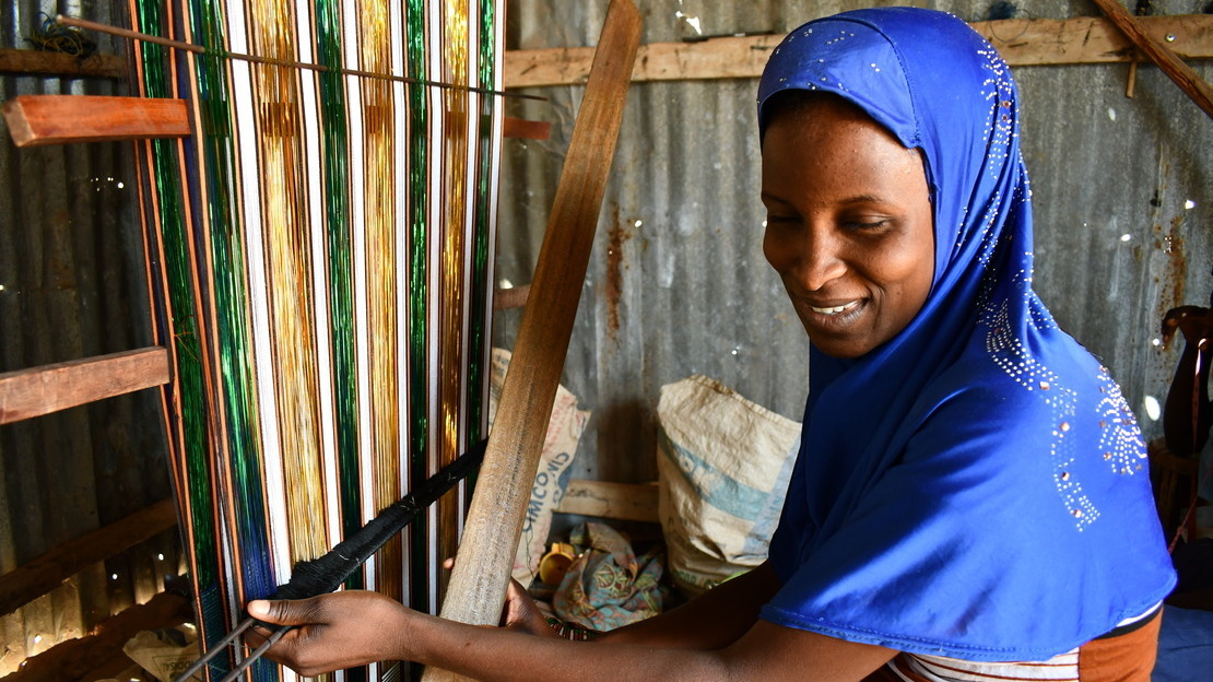 Portrait d'un jeune fille portant un voile bleu, le visage tourné vers la caméra et qui sourit. Elle se tient devant un métier à tisser qu'elle manipule avec ses deux mains.