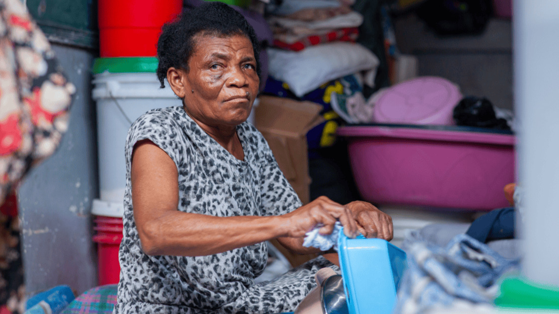 Une femme est assise et nettoie un contenant en plastique à l'aide d'un chiffon. Derrière elle, un empilement de seaux, de tissus, de cartons, etc.