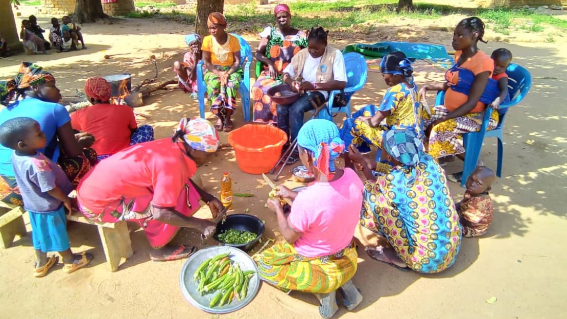 Un groupe de femmes et d'enfants est installé en cercle, sous les arbres, dans le cadre d'une activité de cuisine.