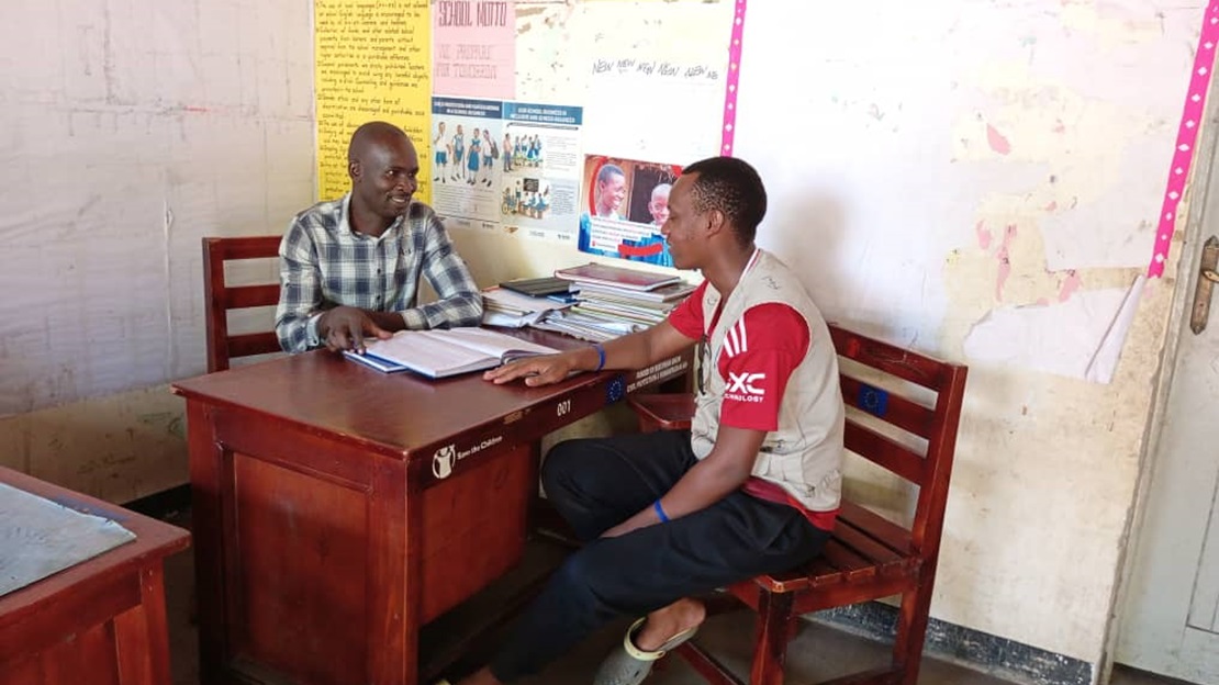 Deux adultes sont en train de discuter dans une salle de classe, de part et d'autre d'un bureau.
