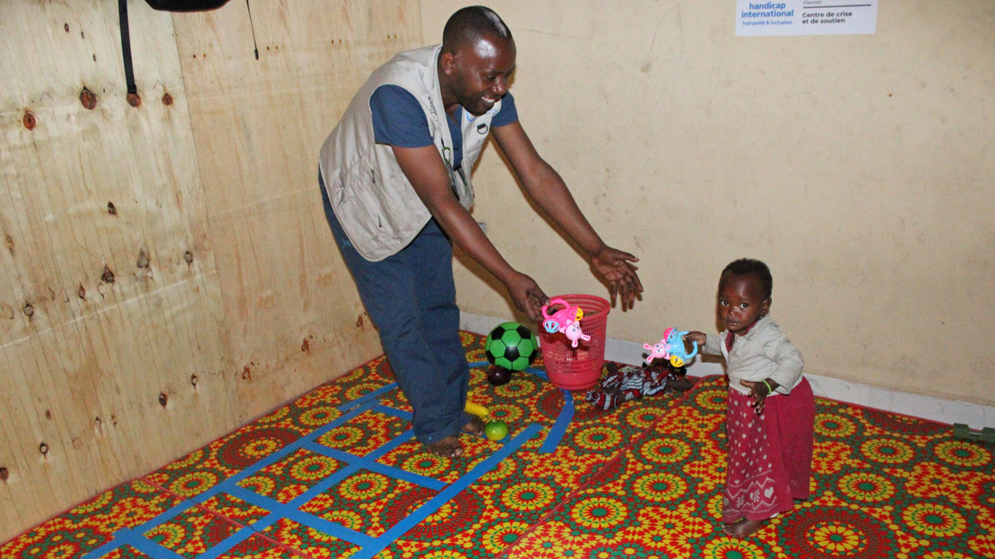 Un kinésithérapeute tend les bras en souriant à une petite fille qui se tient debout devant lui, un jouet en plastique à la main.