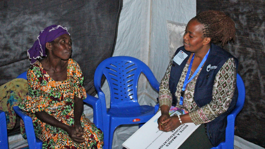 Deux femmes sont assises sur des chaises dans une tente. Elles se regardent et discutent. Celle à droite porte une jaquette floquée HI et tient sur ses genoux un carnet intitulé 