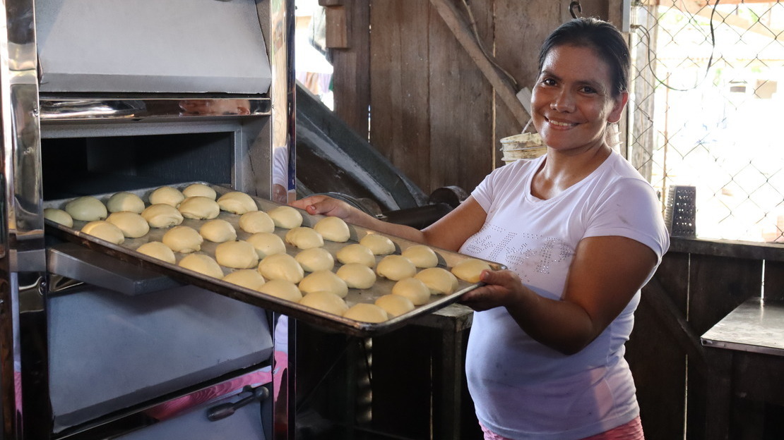 Tatiana tient un plateau couvert de petites boules de pâte devant la porte ouverte d'un grand four. Elle sourit à la caméra. 