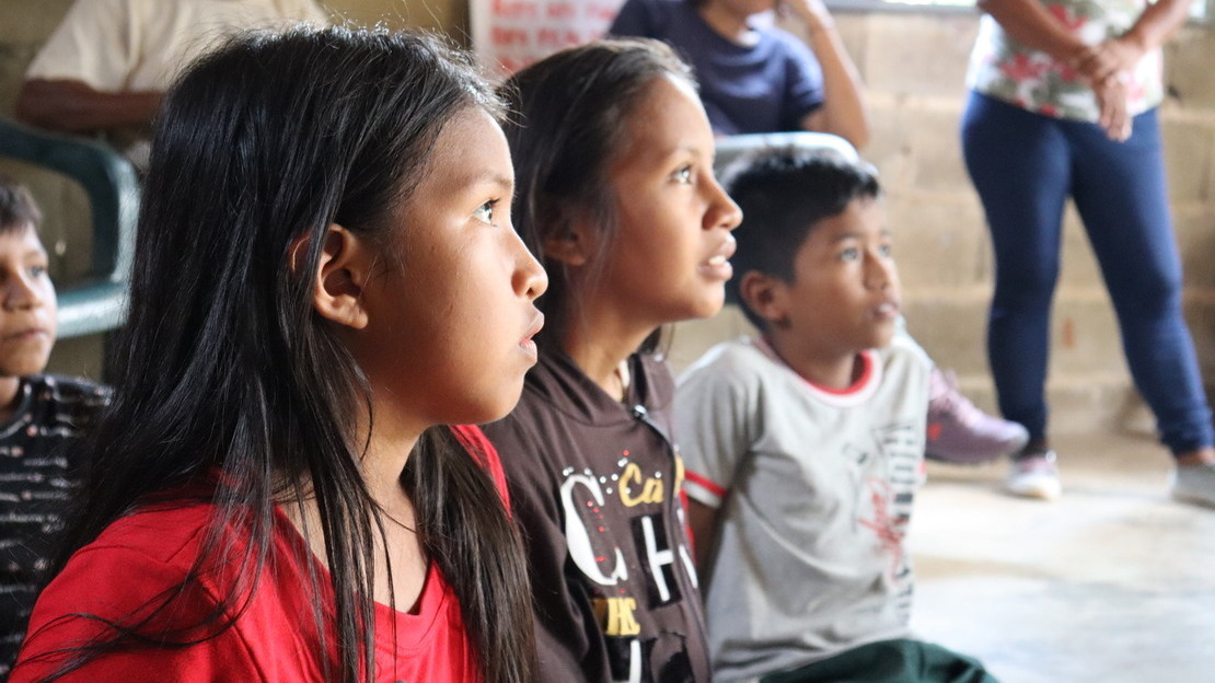 Trois visages d'enfants en gros plan, deux filles et un garçon. Ils sont assis par terre et regardent devant eux. En arrière-plan, on devine des adultes se tenant debout.