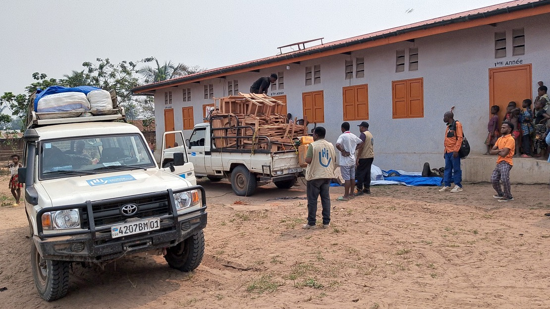 Un véhicule chargé de bancs d'école en bois est garé devant un bâtiment scolaire. Des hommes s'apprêtent à le décharger.