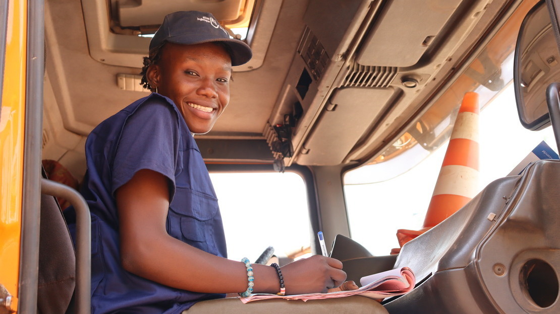 Une jeune femme noire est assise dans une cabine de camion. Elle tient un stylo et des papiers et regarde en souriant le photographe en contrebas.