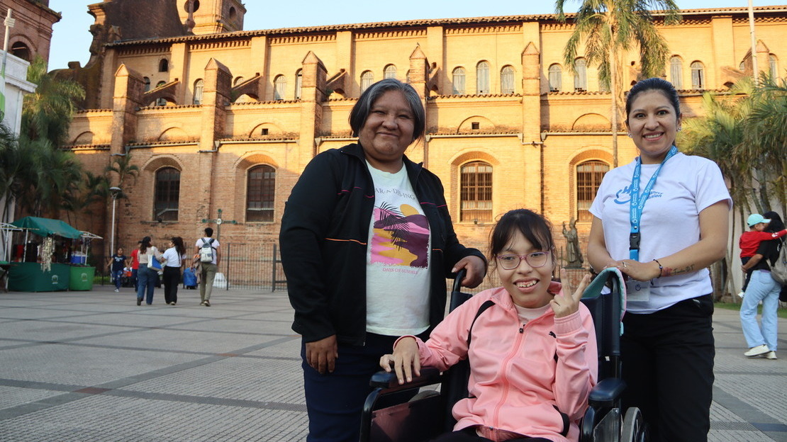 Trois femmes se tiennent sur une place, devant une cathédrale illuminée par le soleil. Deux d'entre elles sont debout et entourent la troisième, assise dans un fauteuil roulant. Elles sourient au photographe.
