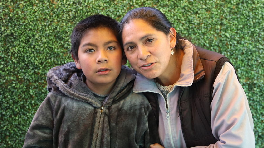 Portait en gros plan d'un garçon et de sa maman, sur fond vert. Ils se serrent tendrement l'un contre l'autre et regardent le photographe en souriant.