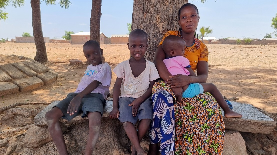 L'image montre une femme et deux enfants assis côte à côte sur un banc pierre au pied d’un grand arbre. La femme porte un enfant dans les bras.