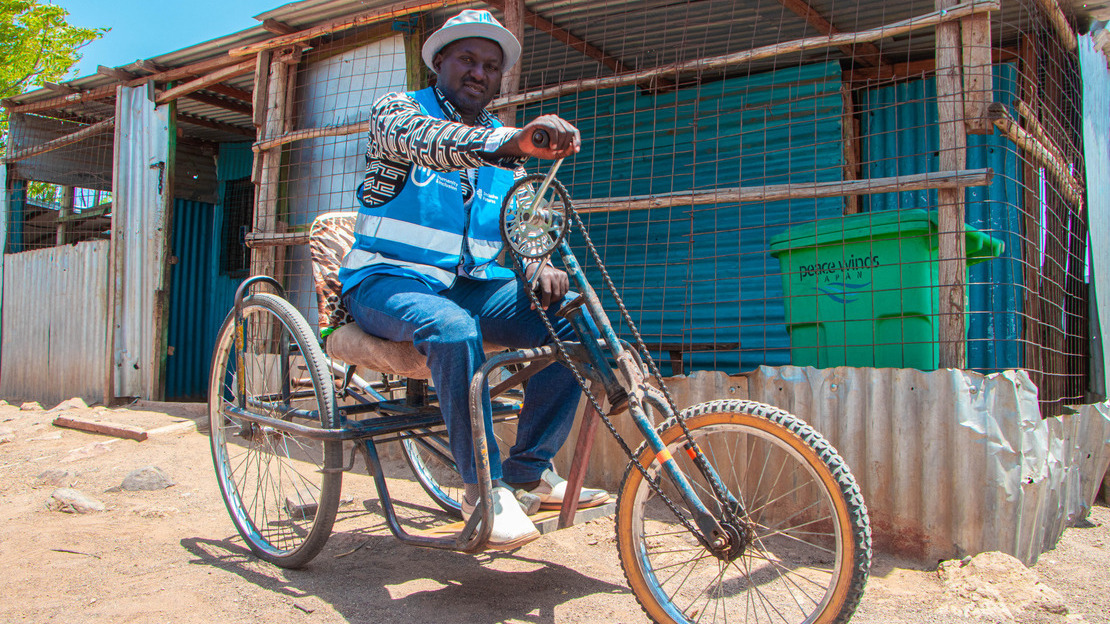 Un homme est assis dans un tricycle dans une rue. Derrière lui on devine une maison en tôle. Il sourit au photographe.