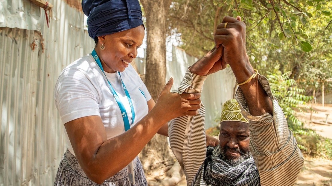 Gros plan sur une femme qui tient les bras levés devant lui d'un homme assis. Ils se trouvent dans une cour ombragée.