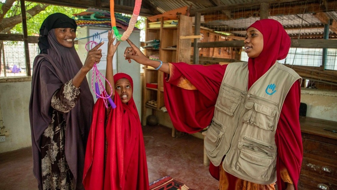 Une femme portant une jaquette HI désigne un ruban qui pend du plafond à une fillette qui tend la main pour l'attraper. Elles sont accompagnée d'une troisième femme, toutes sourient.