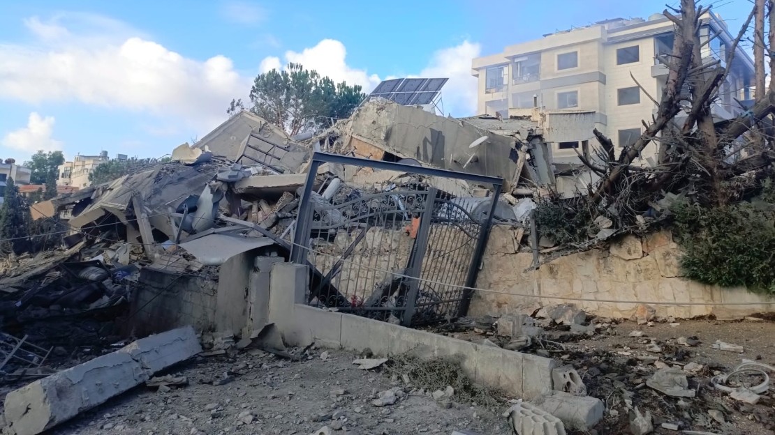 Bâtiment effondré, avec des blocs de béton brisés, une grille métallique tordue et des arbres endommagés au premier plan. En arrière plan, on voit d’autres immeubles intacts sous un ciel nuageux.